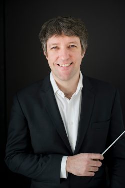 Professional branding portrait of a classical music conductor in a black suit holding a baton against a dark studio backdrop in Milwaukee, Wisconsin.