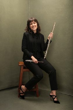Editorial branding portrait of a female classical flutist dressed in black, seated on a wooden stool and holding a silver flute against a muted olive studio backdrop in Milwaukee, Wisconsin.