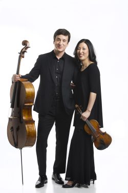 High-key studio branding portrait of a married classical music duo, a male cellist and female violinist, dressed in black and posing together with their instruments against a white backdrop.