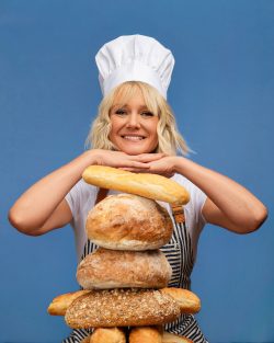 Creative Milwaukee branding portrait of Serai, owner of Whisk & Wonder Cooking Club, wearing a chef's hat and posing playfully behind a stack of artisan breads against a blue studio backdrop.