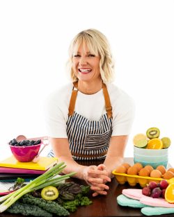 Lifestyle branding portrait of Serai, owner of Whisk & Wonder Cooking Club, smiling in a striped apron behind a colorful spread of fresh fruits and vegetables in a bright studio.