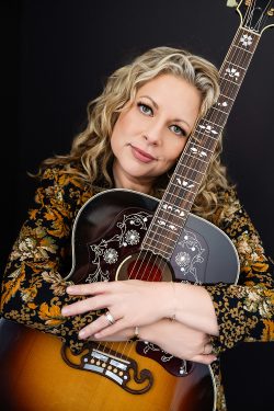 Editorial branding portrait of a blonde female musician embracing an acoustic guitar, photographed against a dark backdrop at Jennifer Brindley Portrait in Milwaukee, Wisconsin.