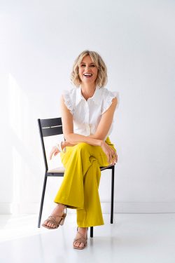 Milwaukee personal branding portrait of the interior designer and owner of Color Zen Interiors, smiling in a white blouse and vibrant yellow trousers while seated on a black chair in a bright studio.