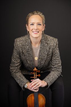 Editorial branding portrait of Margot, a professional violinist and board member, smiling in a textured blazer with her violin resting in front of her against a dark studio backdrop.